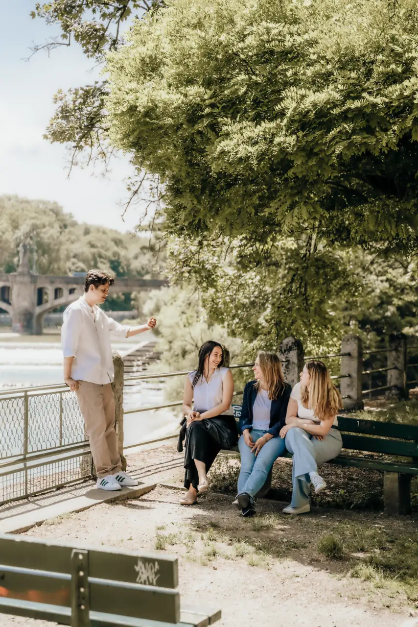 Vier Mitarbeitende von RIFA bei einer Pause auf einer Bank am Isarufer, im Hintergrund Wehr und Steinbrücke